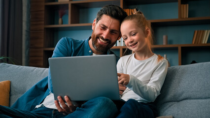 Cheerful father and little beautiful daughter girl sit on couch watch movies cartoons on internet...