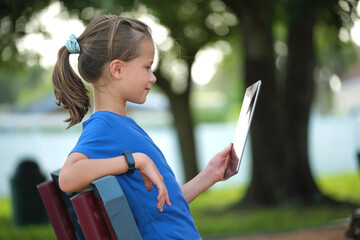 Happy child girl looking at screen of digital tablet resting on bench in park on summer sunny day. Online study concept