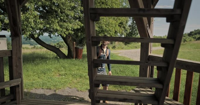 Hiker man with backpack climbing to the top of a wooden tower on stairs in nature using his legs and arms with trees and grass agricultural fields in background in Slovenija, Sotinski breg