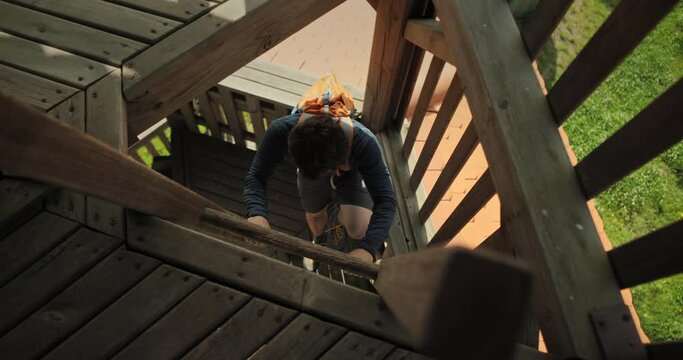 High perspective shot of a hiker man with backpack climbing to the top of a wodden tower on stairs outside using his legs and arms in Slovenija, Sotinski breg