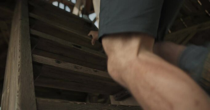 Low perspective shot of a hiker man with backpack climbing to the top of a wodden tower on stairs outside using his legs and arms in Slovenija, Sotinski breg