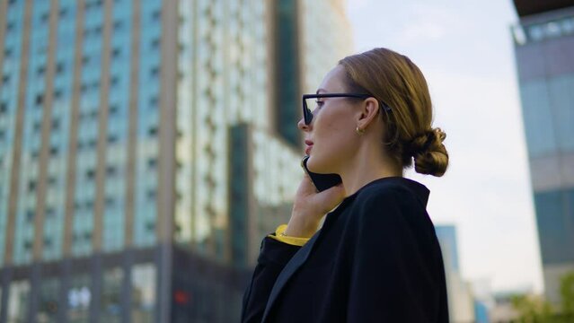 Dissatisfied busy businesswoman in formal outfit and glasses talks on phone negatively shakes head standing at Office skyscraper terrace outside - orbit closeup