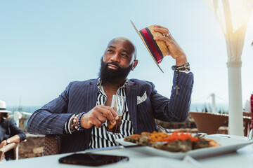 Portrait of a burly elegant bald African guy with a black beard and in a tailored suit, having lunch in a coastal street restaurant, with a glass of beverage in hand lifting his straw hat in greeting