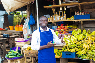 Obraz premium happy african male fruits trader smiling holding a banana with a colorful market fruits background