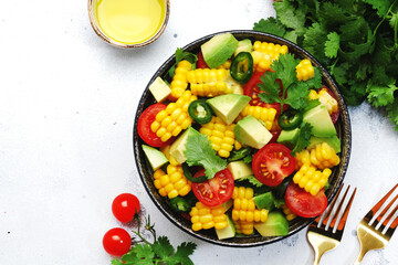 Mexican spicy salad with corn, avocado, jalapeno peppers, cherry tomatoes and cilantro. White kitchen table background, top view
