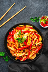 Asian cuisine, stir fry with chicken, red paprika pepper and zucchini in frying pan. Black kitchen table background, top view, copy space
