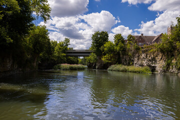 Fototapeta premium Nature landscape - bridge over the rapid Grand River, rocky banks, green trees, cloudy sky, day time. Fergus, Ontario, Canada