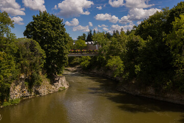 Fototapeta na wymiar Nature landscape - bridge over the rapid Grand River, rocky banks, green trees, cloudy sky, day time. Fergus, Ontario, Canada