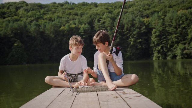 Brothers schoolboys sit on wooden pier and play with bait. Brothers schoolboys sit cross legged on wooden pier near net on sunny summer day. Boys hold rods and baits in hands. Elder boy examines red