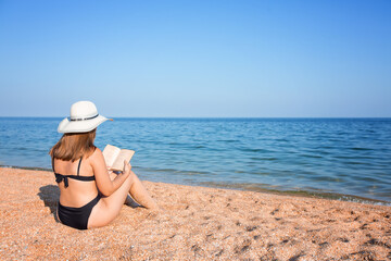 Fototapeta na wymiar An attractive Caucasian girl in a swimsuit and a straw hat sits on the shore and reads an interesting book on the sea horizon. Back view