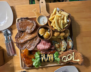 Beef, fish, chicken and pork steaks are laid out on a large wooden tray, served with french fries and mashed potatoes.