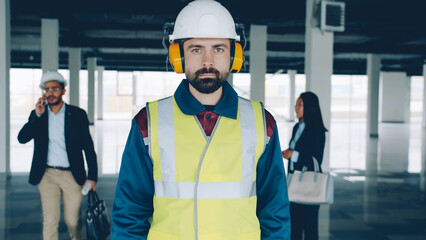 Fototapeta na wymiar Portrait of serious construction specialist wearing safety uniform standing in empty building with businesspeople in background. Real estate and repairs concept.