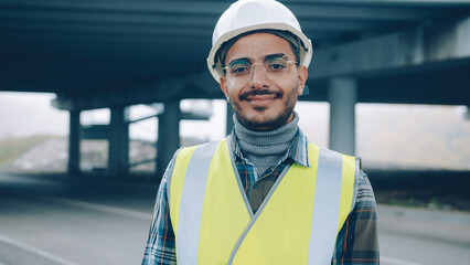 Joyful Arab guy wearing construction uniform is standing in urban building area smiling and looking at camera. People and workplace concept.