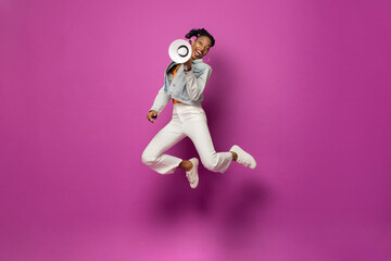 Energetic trendy young African American woman jumping with megaphone in studio purple pink color...