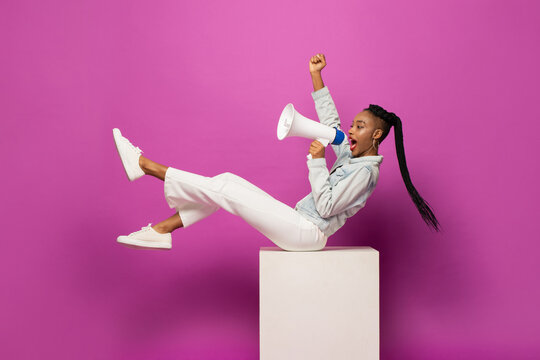 Young African American woman shouting on megaphone and raising fist in studio purple pink color isolated background