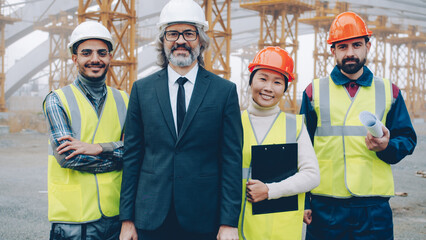 Successful architects multiracial team are standing outdoors at project site smiling holding papers and blueprints. People and occupation concept.