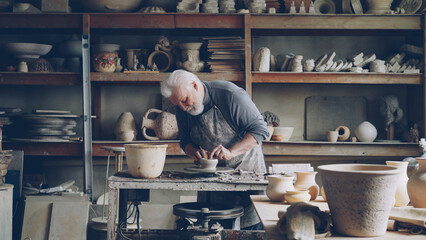 Experienced male ceramist is creating utensils from clay on potter's wheel in workshop. Producing...