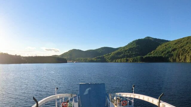 Vista de un lago desde un ferry que est&aacute; moviendose antes de llegar a puerto. Se ve el agua, monta&ntilde;as de bosques verdes y cielo azul despejado.