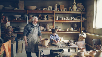 Young grandson is molding clayware in his grandfathers's home studio while his loving senior grandpa is standing near him and watching him with love. Family traditions concept.