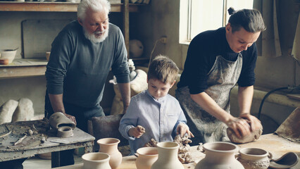 Professional male potter is kneading clay on worktable in home studio while his son is helping him and his elderly father watching them from behind. Small family business concept. - 536890834