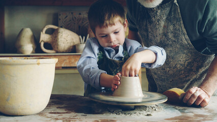 Curious little boy is learning pottery from his experienced grandfather in small home studio. Child is forming clay to make pot on potter's wheel, his granddad is helping him.