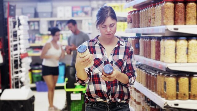 Interested young woman standing near shelves with jars of canned vegetables in supermarket, choosing beans in sauce 