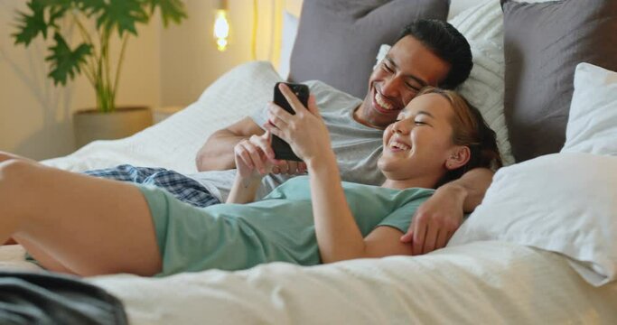 Asian couple, phone and social media for communication and entertainment on bed to relax, laughing and watch funny videp in bedroom at home. Man and woman using 5g internet while lying together