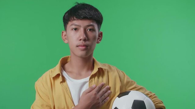 Close Up Of Asian Boy With A Ball Putting Hand On His Heart To Repect The National Flag And Anthem While Cheering Soccer On Green Screen Background
