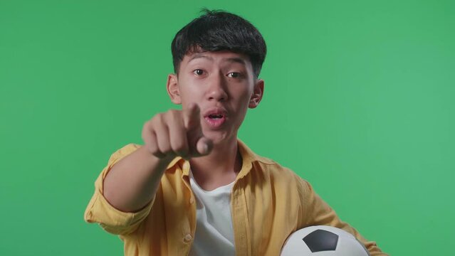 Close Up Of Asian Boy With A Ball Pointing To Camera For Making Fun Of The Losers While Cheering Soccer On Green Screen Background
