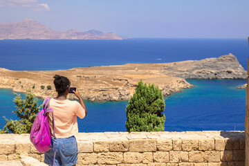 Happy girl enjoys view of coast of ancient old town of city Lindos, Greece. Woman in taking photo...