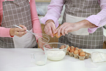 Happy young couple multiethnic man and woman hands making homemade bakery cake, pouring milk while breaking eggs and mixing flour into glass bowl in the kitchen. Family having fun while prepare food