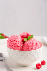 Scoops of sweet organic dairy berry ice cream or refreshing sorbet of pink color decorated with juicy raspberries and fresh mint leaf served in bowl on white wooden table with metal spoon. Copy space