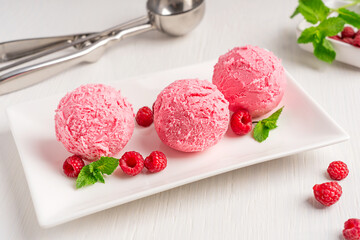 Three scoops of sweet homemade berry ice cream or refreshing sorbet of pink color decorated with juicy raspberries and fresh mint leaf served on plate on white wooden table with metal scooper utensil