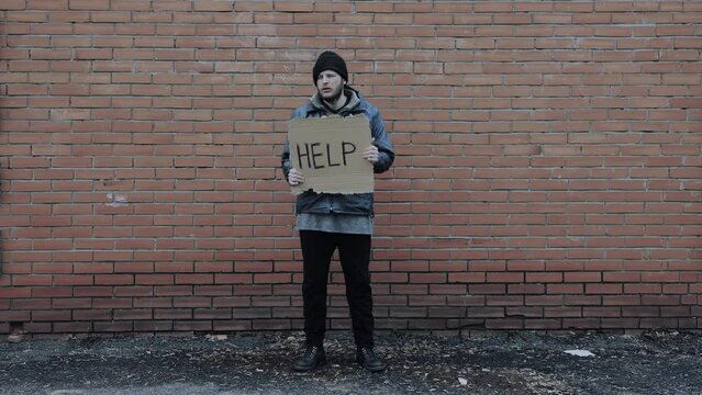 Homeless holds piece of cardboard with inscription Help. Refugee is at brick wall looking for job. Male tramp in dirty clothes. Below poverty line. Bearded fugitive beggar. Immigrant. Drunkenness