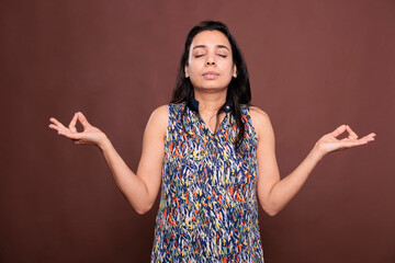 Serene indian woman meditating, showing om sign with fingers, front view studio mid shot on brown background. Relaxed person with closed eyes, calm lady with yoga symbol, meditation gesture - obrazy, fototapety, plakaty