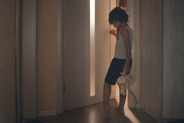 A boy with a toy teddy bear opens the door to a room with light in a dark corridor. Boy in pajamas concept of going to bed. soft focus, toned photo.