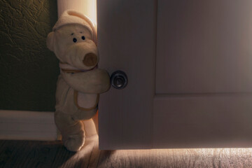 a teddy bear walks out of a room with the lights on into a dark corridor, holding onto a white...