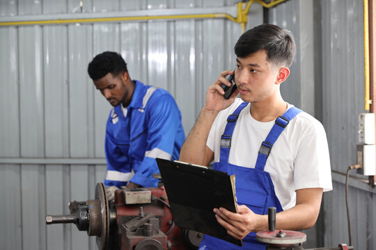 Multiethnic technician mechanic using metal lathe machine operate polishing car disc brake with smart mobile phone and clipboard at garage. Maintenance automotive and inspecting vehicle part concept