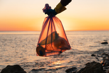 Fototapeta na wymiar Earth Day. Garbage collection on the shore. A volunteer holds a garbage bag with plastic bottles against the background of sunset in the ocean, close-up. The concept of environmental pollution