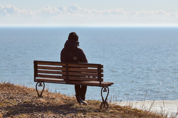 Single girl in a black jacket and hat sitting on bench at cliff at front of sea, peaceful and quiet...