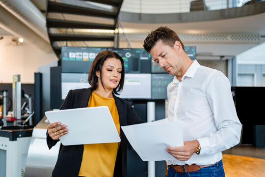 Businesswoman holding tablet PC examining paper standing by colleague in industry