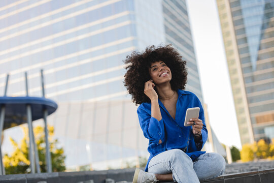 Smiling woman with Afro hairstyle holding mobile phone sitting on wall
