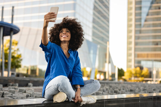 Happy Afro woman talking selfie through mobile phone sitting on wall
