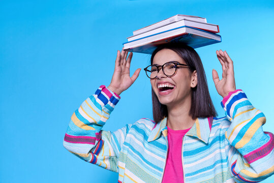 Happy young woman wearing colorful zipper balancing books on head against blue background