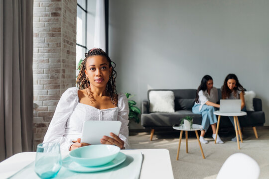 Young Woman Holding Tablet PC With Flatmates In Background At Home