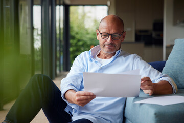Senior man reading documents sitting by sofa at home