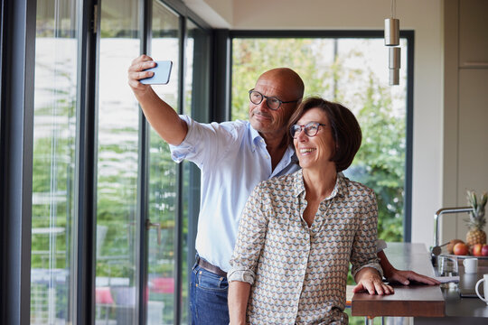 Happy senior woman with man taking selfie through smart phone at home
