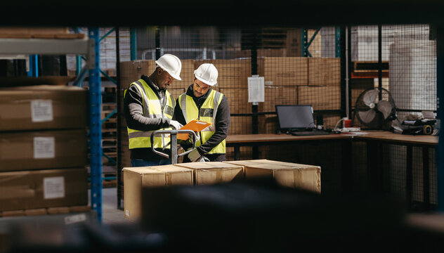 Two warehouse employees reading a clipboard while moving goods with a pallet truck in a logistics center