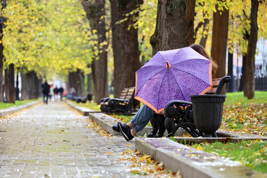 Rain in autumn season, girl with umbrella sitting on a bench in city park on walking people background