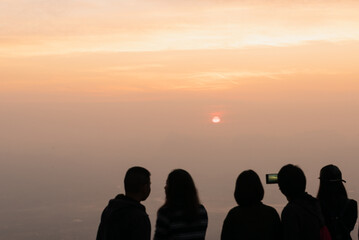 Fototapeta premium Silhouette traveler taking a photo the sun with clouds and relaxing action on top mountain with sunrise on very mist and orange sky background in the morning time.Happy and funny time with friends.